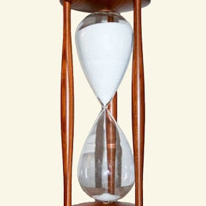 May include: A wooden hourglass with a clear glass bulb filled with white sand. The hourglass is standing on a white surface.