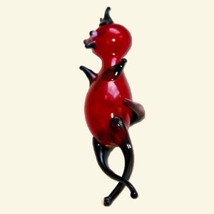 May include: A red glass figurine of a devil with black horns and tail. The figurine is standing on its hind legs with its arms raised.