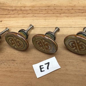May include: Six round ceramic cabinet knobs with a glossy brown and sage green glaze featuring a swirling geometric pattern. Each knob has a silver-toned metal post and base. A small white rectangular paper tag with the black text E7 is included.