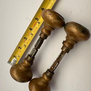 May include: Two antique bronze door knobs with a tarnished finish. The knobs are oval-shaped and connected to a metal rod. A yellow tape measure is in the background, showing measurements in inches.