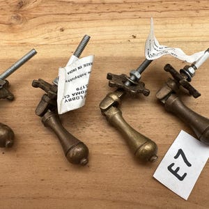 May include: Four brass-toned metal drop pulls with a swinging handle design. Each pull features a decorative star-shaped backplate, a threaded bolt, and a nut. Two pulls have white paper tags attached, and one white paper tag displays the text E7.