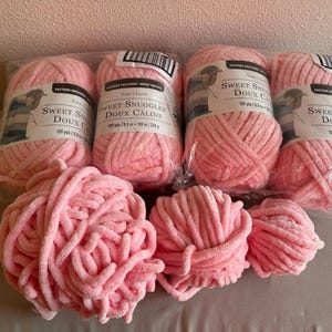 以下が含まれることがあります： 「Sweet Snuggles Doux Calins」の文字が入った、柔らかいピンク色の毛糸の束。一部は包装されたままで、手編みや編み物プロジェクトに最適な、ふわふわとしたベルベットのような質感です。