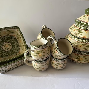 May include: A ceramic dinnerware set featuring a cream base with green leaf patterns, small yellow flowers, and dark green decorative borders. The set includes two square bowls, four mugs, and a three-tiered stackable serving dish with a lid.