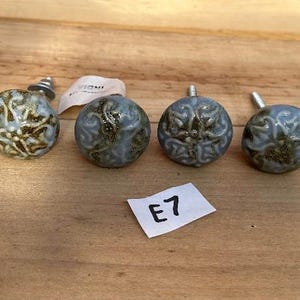 May include: A set of eight round ceramic cabinet knobs featuring a light blue and sage green glazed finish with a raised, intricate embossed pattern. Each knob has a metal bolt extending from the back. A small white paper tag labeled E7 is included.