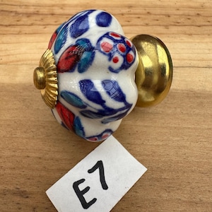 May include: A ceramic drawer knob with a white base featuring a hand-painted floral pattern in blue, red, and teal. The knob has a scalloped, pumpkin-like shape with a gold-toned metal base and a small gold-toned cap. A white paper tag with the text E7 is visible.