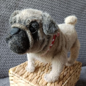 Può includere: Figurina di carlino di peluche con pelo beige e grigio, naso nero e collare rosso a pois bianchi. Il carlino è in piedi su un cestino intrecciato. Il pelo del cane è morbido e dettagliato, con un aspetto realistico.