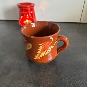 Puede incluir: Una taza de cerámica marrón hecha a mano con un asa, adornada con diseños florales pintados en rojo y dorado. Un pequeño portavelas rojo con una vela encendida está en el fondo. La taza está sobre una superficie oscura.