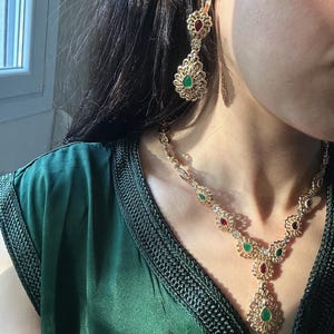 Peut inclure: Ensemble de bijoux dorés comprenant un collier et des boucles d'oreilles. Le collier et les boucles d'oreilles présentent des motifs en filigrane complexes, ornés de pierres précieuses rouges et vertes. Les bijoux sont présentés sur un vêtement vert foncé.