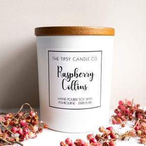 May include: A white candle with a wooden lid, labelled "Raspberry Collins" from The Tipsy Candle Co. The label also states "Hand Poured Soy Wax, Ashbourne, Derbyshire." Pink berries are scattered around the base of the candle.