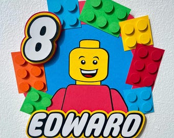 May include: A colorful cake topper featuring a yellow Lego figure with a red shirt, surrounded by Lego-style blocks in various colors. The number "8" and the name "EDWARD" are also included, perfect for a birthday celebration.
