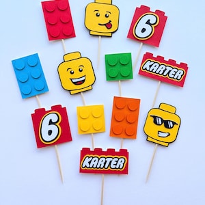 Può includere: Un set di dodici decorazioni per cupcake su stecchini di legno, con teste di minifigure gialle dalle varie espressioni, mattoncini da costruzione rossi, blu, verdi, arancioni e gialli, il numero 6 e il nome KARTER in stampatello.
