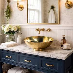 Peut inclure: Meuble-lavabo de salle de bain luxueux avec un lavabo doré, une robinetterie dorée et un comptoir en marbre. Le meuble bleu marine comporte trois tiroirs et deux paniers en osier. Un grand miroir doré et des appliques murales complètent le design. Le meuble mesure 120 cm de large.
