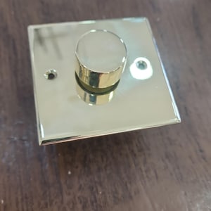May include: A gold dimmer switch with a square base and a cylindrical knob. The switch has a polished, reflective surface and two small holes. The dimmer is designed for electrical use.