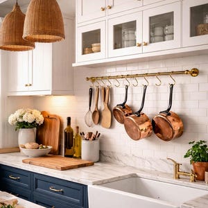 Könnte beinhalten: Eine Küche mit gewebten Pendelleuchten, weißen Schränken und einer Marmorarbeitsplatte. Kupferpfannen hängen über einem weißen Bauernhauswaschbecken. Holzschneidebretter und Utensilien verleihen Wärme. Die Schränke sind marineblau.