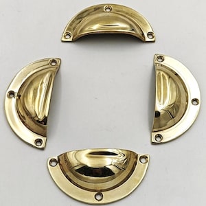 May include: Four polished brass half-moon drawer pulls. Each pull has a curved design with three screw holes for mounting. The pulls are a bright gold color and reflect light.