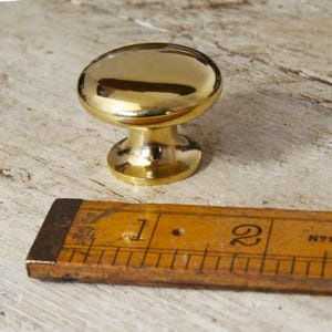 Op de afbeelding: Een glanzende, goudkleurige kastknop met een afgeronde bovenkant en een korte voet. Een vintage houten liniaal met centimeter markeringen is gedeeltelijk zichtbaar. De knop is waarschijnlijk voor meubels of kasten.