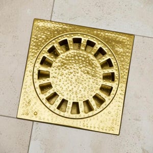 Könnte beinhalten: Ein quadratischer, goldfarbener Bodenablauf mit strukturierter Oberfläche und einem kreisförmigen, geschlitzten Design in der Mitte. Der Ablauf besteht aus Metall und ist für den Einbau in einen gefliesten Boden konzipiert. Der Ablauf ist ein dekoratives Element für ein Badezimmer oder eine Dusche.