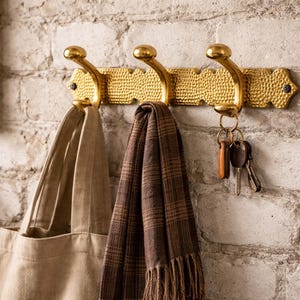 May include: A gold-colored metal wall hook with three curved hooks. A beige tote bag, a brown plaid scarf, and a set of keys hang from the hooks. The wall is made of white bricks.
