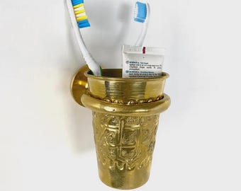 May include: A gold-colored toothbrush holder with two toothbrushes and a tube of toothpaste. The holder is mounted on a white wall. The toothbrushes have white handles and blue and yellow bristles.