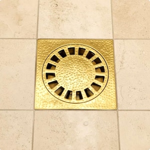 Peut inclure: Un couvercle de drain de douche carré doré avec une surface texturée et un design circulaire à fentes. Le couvercle de drain est encastré dans un sol carrelé beige.