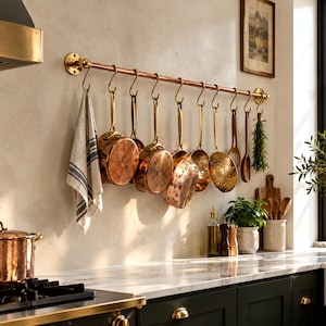 May include: A wall-mounted copper rail with brass end brackets features ten S-hooks holding a striped kitchen towel, various copper pots and pans, a perforated skimmer, wooden utensils, and a bundle of dried herbs in a kitchen.