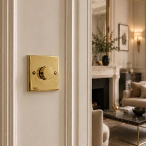 May include: A square, polished brass light dimmer switch mounted on a white wall with vertical moulding. The switch features a central circular knob and two flat-head screws on the left and right sides of the metallic faceplate, reflecting light.