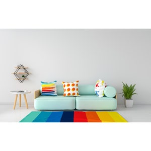 May include: A light blue sofa with three colorful throw pillows. The sofa is in front of a white wall with a geometric shelf above it. A small wooden table with a white top sits in front of the sofa. A rainbow striped rug is on the floor in front of the sofa.