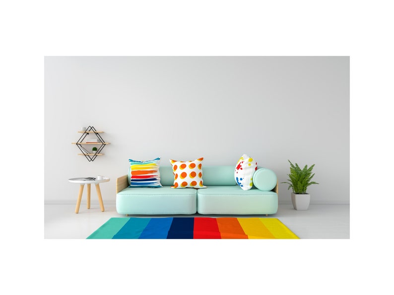 May include: A light blue sofa with three colorful throw pillows. The sofa is in front of a white wall with a geometric shelf above it. A small wooden table with a white top sits in front of the sofa. A rainbow striped rug is on the floor in front of the sofa.