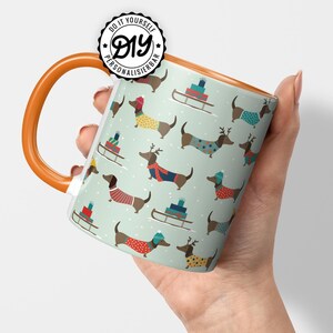 May include: A ceramic mug with an orange handle and rim, featuring a repeating pattern of dachshunds in sweaters and hats, with sleds and gifts on a light green background. The mug has the text "DIY PERSONALISIERBAR" on it.