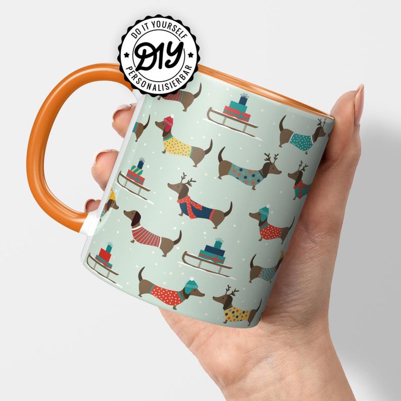 May include: A ceramic mug with an orange handle and rim, featuring a repeating pattern of dachshunds in sweaters and hats, with sleds and gifts on a light green background. The mug has the text "DIY PERSONALISIERBAR" on it.