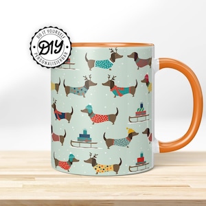 May include: A ceramic mug with an orange handle and interior. The exterior features a repeating pattern of dachshunds wearing sweaters, hats, and antlers, with snowflakes and sleds on a light green background. The mug has the text "DIY PERSONALISIERBAR".