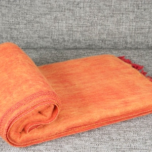 Op de afbeelding: Een oranje deken met rode franjes, opgerold. De deken is gemaakt van een zacht, pluche materiaal.