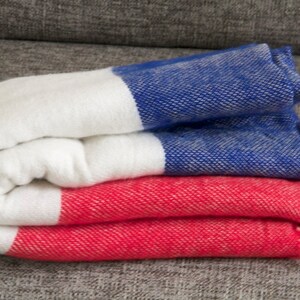 May include: Folded textile with a color block design in white, blue, and red. The woven fabric has a textured appearance and is neatly stacked. The ends of the textile have a fringe detail. The textile is likely a blanket or throw.