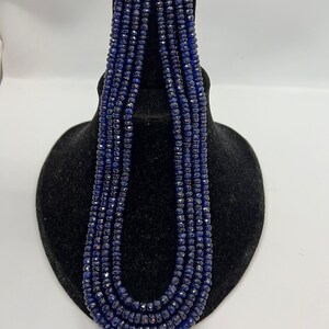 Può includere: Una collana di perle di zaffiro blu scuro, esposta su un supporto in velluto nero. Le perle sono sfaccettate e hanno una forma leggermente irregolare.