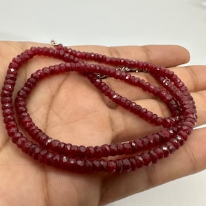 May include: A deep red beaded necklace with a faceted design. The necklace is composed of small, evenly sized beads, and is secured with a silver clasp. The beads have a polished, reflective surface, and the necklace is displayed on a light background.