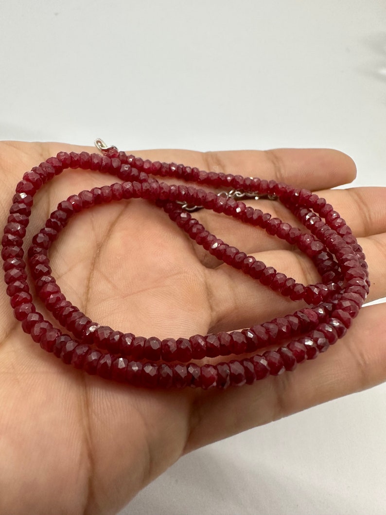 May include: A deep red beaded necklace with a faceted design. The necklace is composed of small, evenly sized beads, and is secured with a silver clasp. The beads have a polished, reflective surface, and the necklace is displayed on a light background.