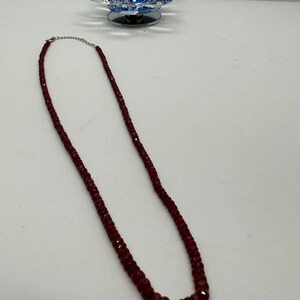 May include: A burgundy-colored beaded necklace with a silver clasp. The necklace is composed of small, faceted beads. The necklace is displayed on a white surface, with a decorative glass lotus flower in the background.