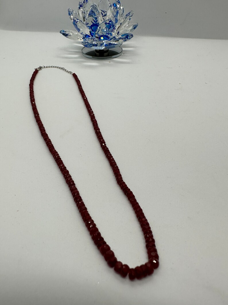 May include: A burgundy-colored beaded necklace with a silver clasp. The necklace is composed of small, faceted beads. The necklace is displayed on a white surface, with a decorative glass lotus flower in the background.