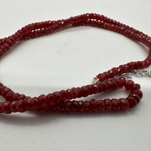 May include: A faceted, deep red gemstone necklace. The necklace is composed of small, evenly sized beads strung together. The clasp is silver-toned. The beads have a sparkling, polished appearance, reflecting light. The necklace is displayed against a white background.
