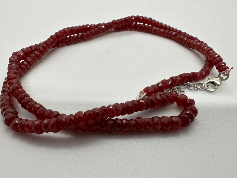 May include: A faceted, deep red gemstone necklace. The necklace is composed of small, evenly sized beads strung together. The clasp is silver-toned. The beads have a sparkling, polished appearance, reflecting light. The necklace is displayed against a white background.