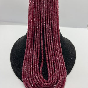 May include: A multi-strand necklace of small, faceted, deep red beads. The beads are strung together, creating a cascading effect. The top of the necklace has a gold-coloured woven detail. The necklace is displayed on a black stand against a white background.