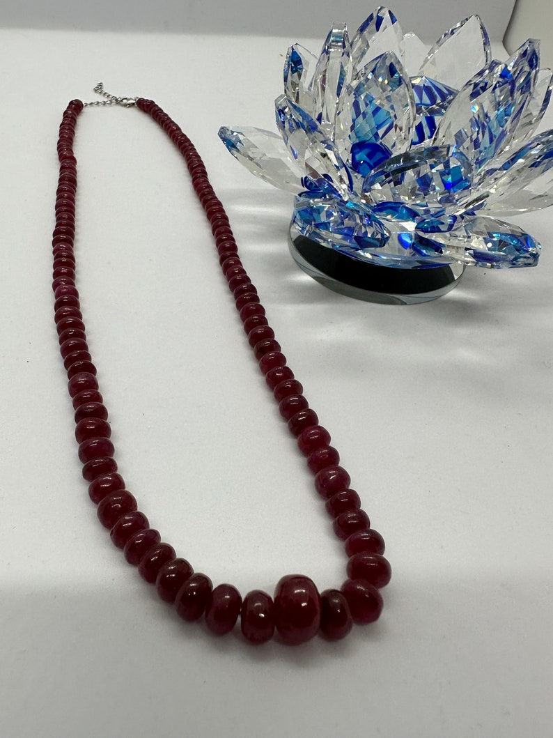 May include: A long necklace made of dark red beads. The necklace has a silver clasp and a larger, round bead in the center.