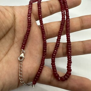 May include: A burgundy-colored necklace composed of small, faceted beads. The necklace is held in a hand, showcasing its length and the silver-toned clasp. The beads appear to be evenly sized and strung together, creating a delicate jewelry piece.