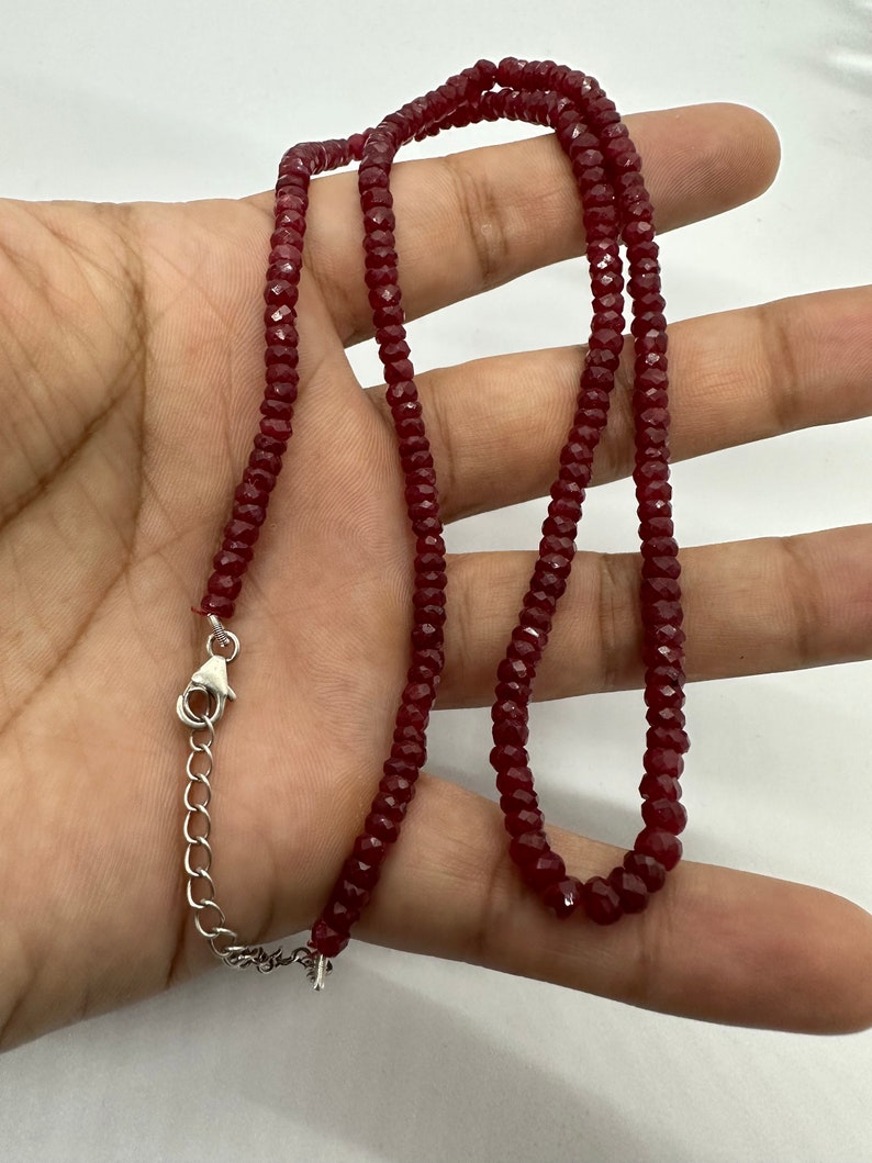 May include: A burgundy-colored necklace composed of small, faceted beads. The necklace is held in a hand, showcasing its length and the silver-toned clasp. The beads appear to be evenly sized and strung together, creating a delicate jewelry piece.