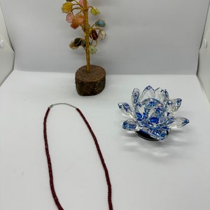 Op de afbeelding: Een bordeauxrode kralenketting met een zilveren sluiting ligt op een wit oppervlak. Een decoratieve kristallen lotusbloem met blauwe accenten en een kleine edelsteenboom zijn ook aanwezig. De boom heeft een houten basis en gouden draad takken.