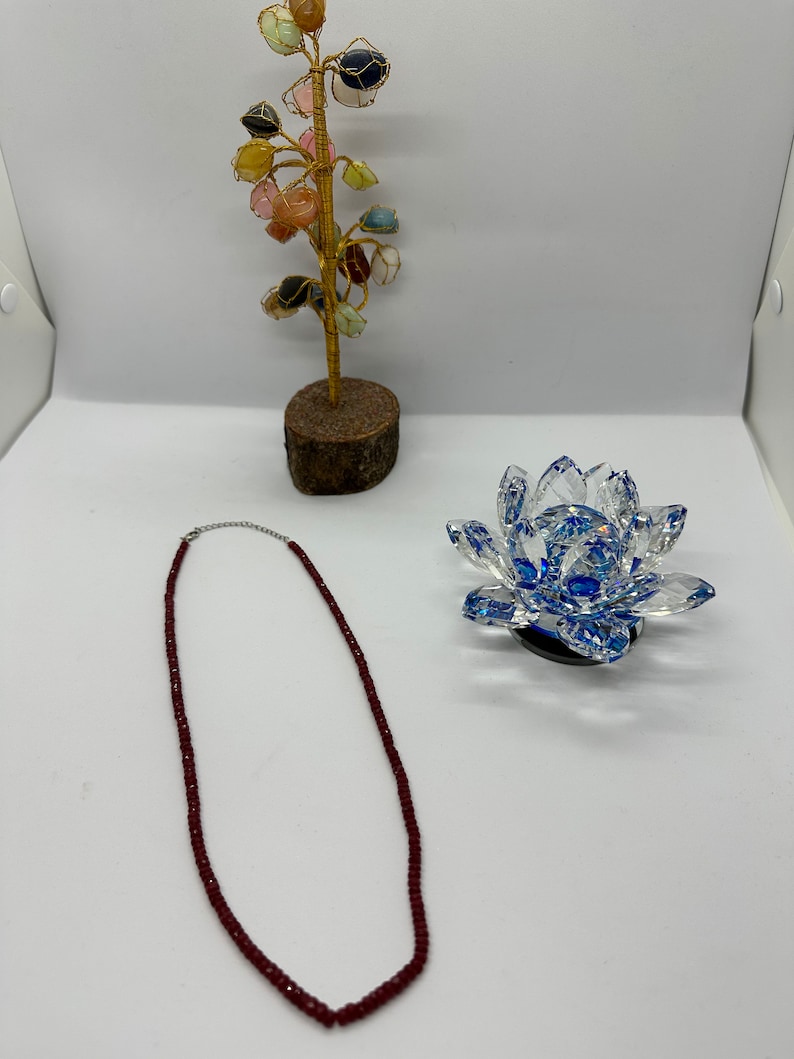 May include: A burgundy beaded necklace with a silver clasp lies on a white surface. A decorative crystal lotus flower with blue accents and a small gemstone tree are also present. The tree has a wooden base and gold wire branches.