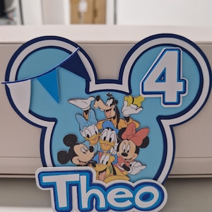May include: A blue and white Mickey Mouse-themed cake topper for a fourth birthday. The design features Mickey, Minnie, Donald, Daisy, Goofy, and Pluto. The name "Theo" is displayed at the bottom, with a small banner.