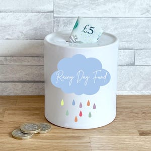 May include: A white ceramic money box with a blue cloud design and the text "Rainy Day Fund". It features colorful raindrop graphics. A five-pound note is in the slot, with coins nearby.