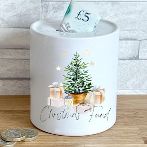 May include: White ceramic money bank with a Christmas tree, presents, and star design. A five-pound note is visible in the slot. The words "Christmas Fund" are written in cursive.