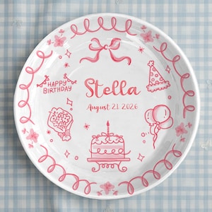 May include: White ceramic plate with a pink birthday theme. The plate features the name "Stella", the date "August 21 2026", and illustrations of a cake, balloons, and flowers. The rim is decorated with pink swirls and floral accents.